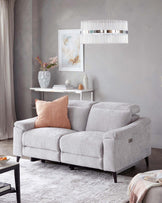 Contemporary light grey fabric upholstered three-seater sofa with plush seat and back cushions, accented with one coral pink throw pillow. The sofa features rounded armrests and dark wooden legs, with a recliner function button visible on the side. A white metal console table with a lower shelf is behind it, adorned with decorative vases, books, and a framed abstract painting hung above. A modern pendant light with vertical fringing and metallic accents is suspended from the ceiling.