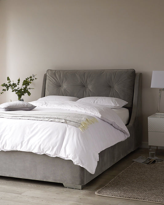 A plush upholstered king-size bed with a tufted headboard in a muted grey fabric, accompanied by a white and grey chevron-patterned nightstand complemented by a simple white lamp. A neutral-toned braided area rug is partially visible at the foot of the bed, adding texture to the serene bedroom setting.