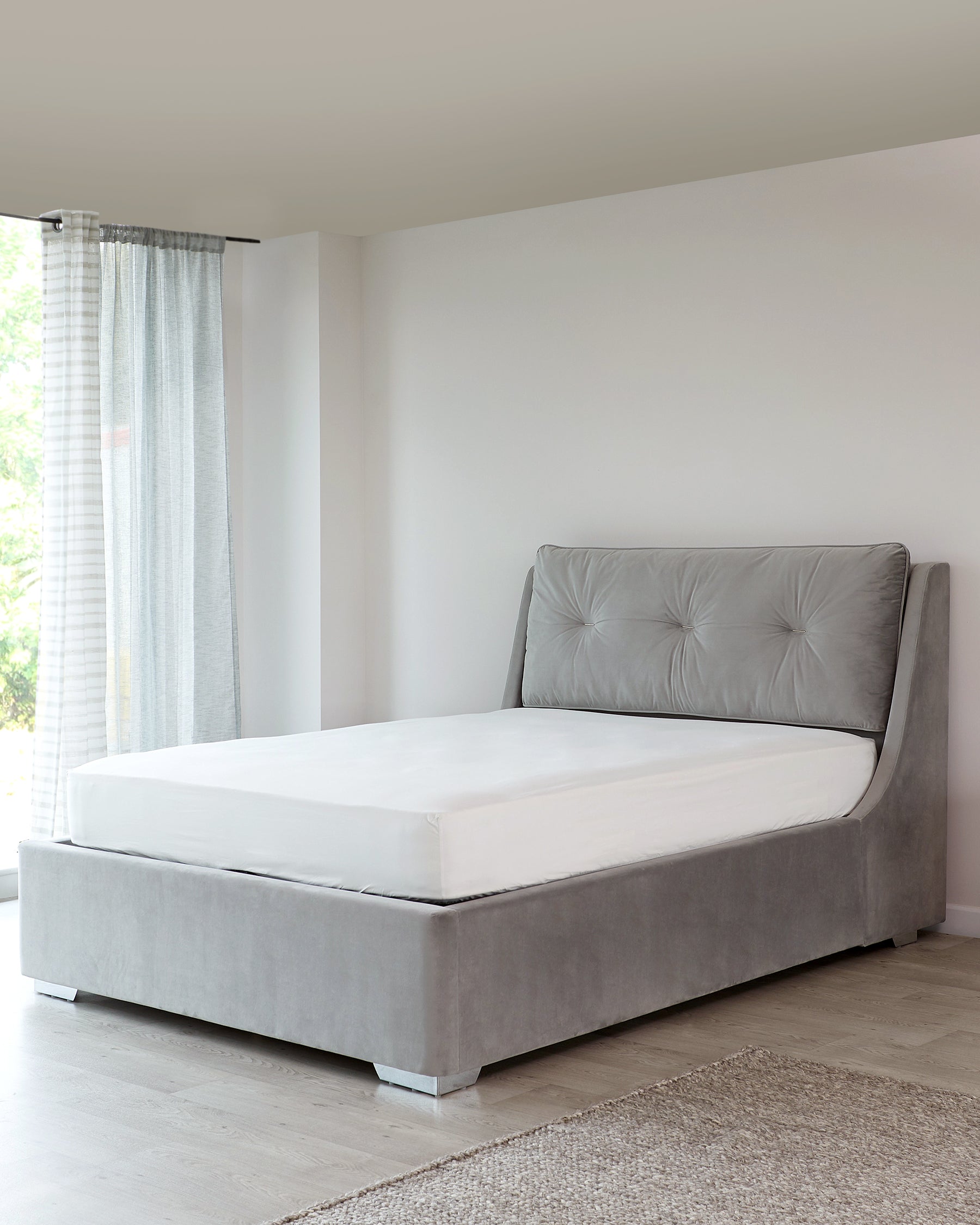 Modern grey upholstered platform bed with button-tufted headboard, featuring clean lines and minimalist design, set against a neutral-toned room with sheer curtains and a textured rug.