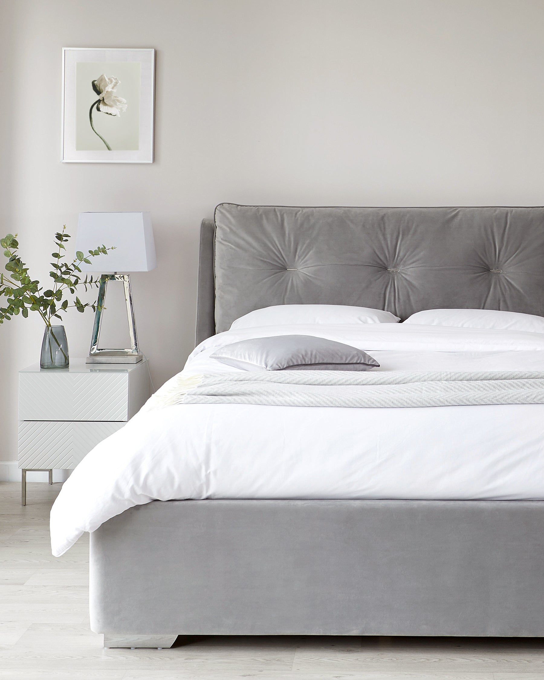 Elegant grey upholstered bed with a tufted headboard and a pristine white bedding set, accompanied by a sleek white bedside table featuring clear glass lamp with white lampshade.