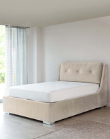 Elegant upholstered platform bed with a high tufted headboard and sleek, modern frame covered in a beige fabric, featuring sturdy low-profile metallic legs. A simple white mattress sits atop the bed frame, and a lush beige area rug is partially visible beneath the bed.