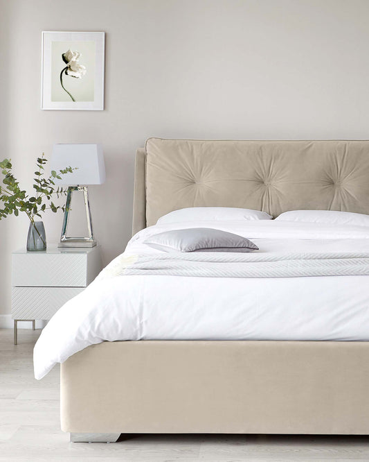 A modern minimalist bedroom featuring a beige upholstered bed with a tufted headboard and a white bedside table with a geometric design, showcasing a clear glass table lamp with a white lampshade. A white framed artwork hangs above the bedside table, complementing the overall clean and tranquil aesthetic.