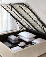 Beige upholstered storage bed with a lifted mattress platform revealing a spacious storage compartment containing pillows, blankets, and a pair of shoes. The bed features a gas-lift mechanism with black metal framing supporting wooden slats under the mattress.