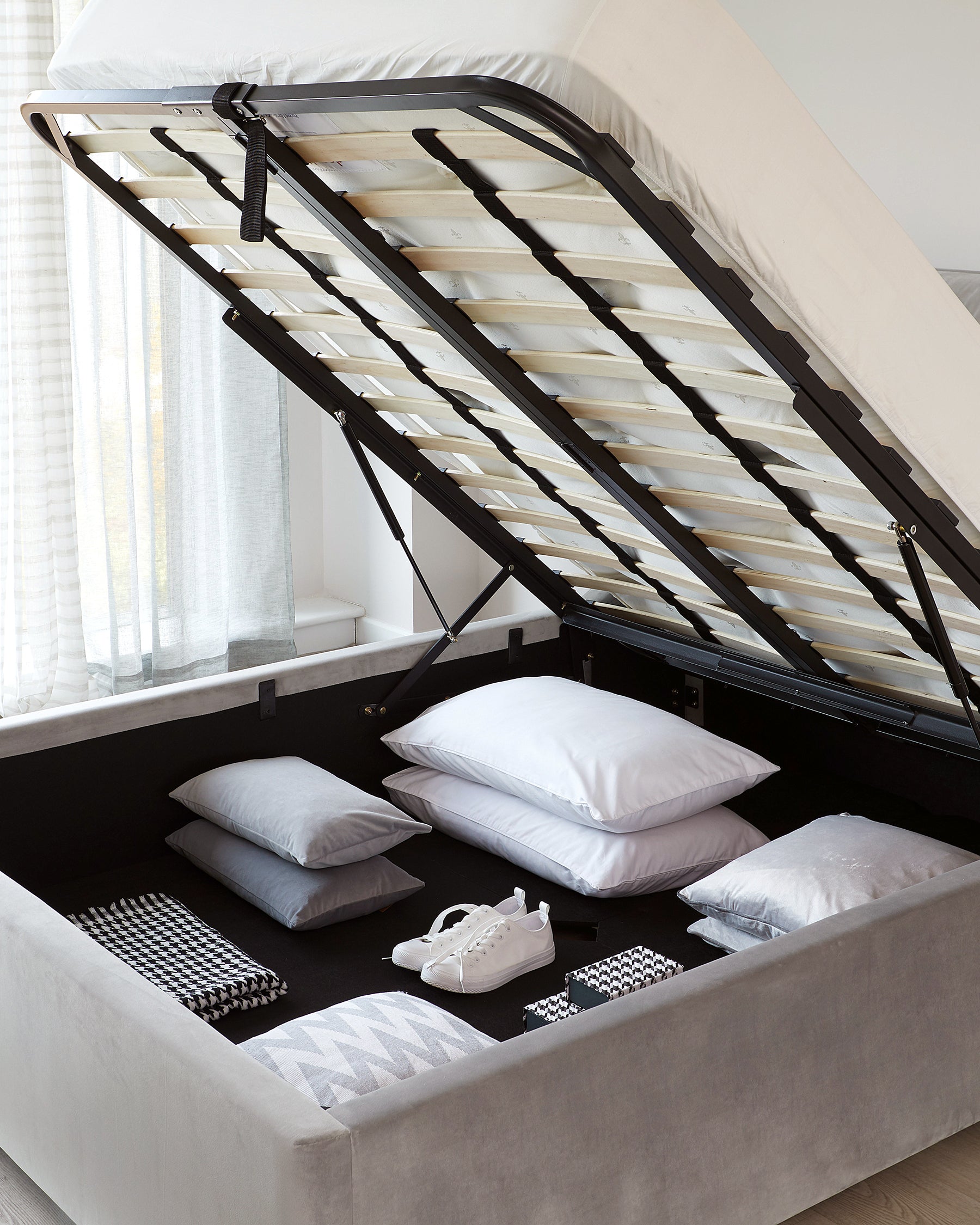 A contemporary grey upholstered storage bed with a lifted frame, revealing a spacious storage area beneath. The bed features a slatted base and is accessorized with various pillows and a pair of white sneakers.