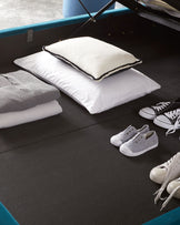 A sleek, modern storage bench with a hinged blue upholstered lid partially lifted, revealing a spacious compartment inside. The bench is positioned on a dark-coloured carpeted floor and accessorized with a stack of casual pillows and an arrangement of sneakers nearby, suggesting a relaxed, contemporary lifestyle vibe.