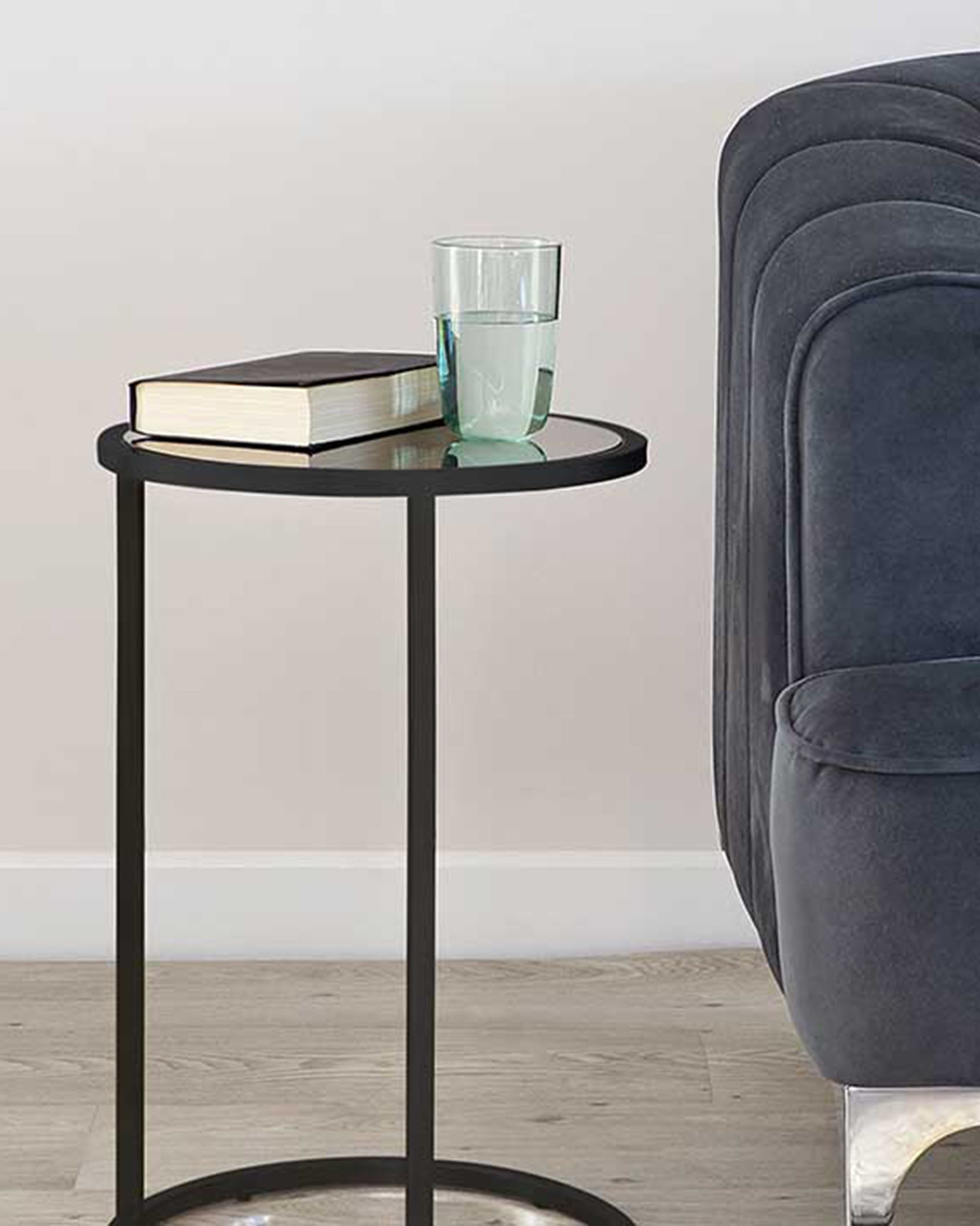 A contemporary round side table with a matte black metal frame and a sleek black tabletop, displayed next to the arm of a plush grey upholstered chair with a book and a glass of water resting on the table surface.