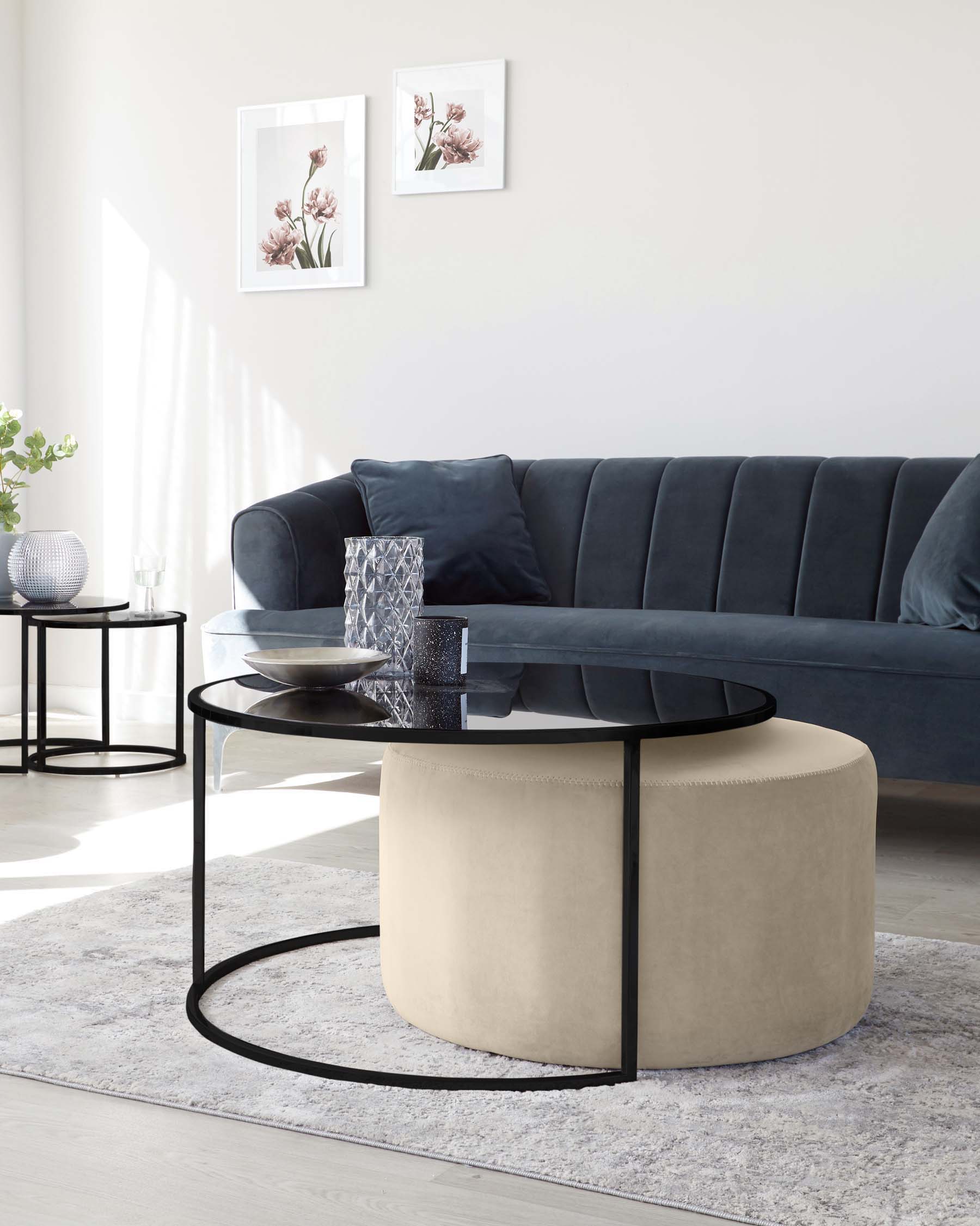 Elegant modern living room furniture featuring a dark grey upholstered sofa with tufted backrest, a round two-tiered coffee table with a black frame and glass top, and a round, light beige ottoman with a tailored, stitch-detailed finish.