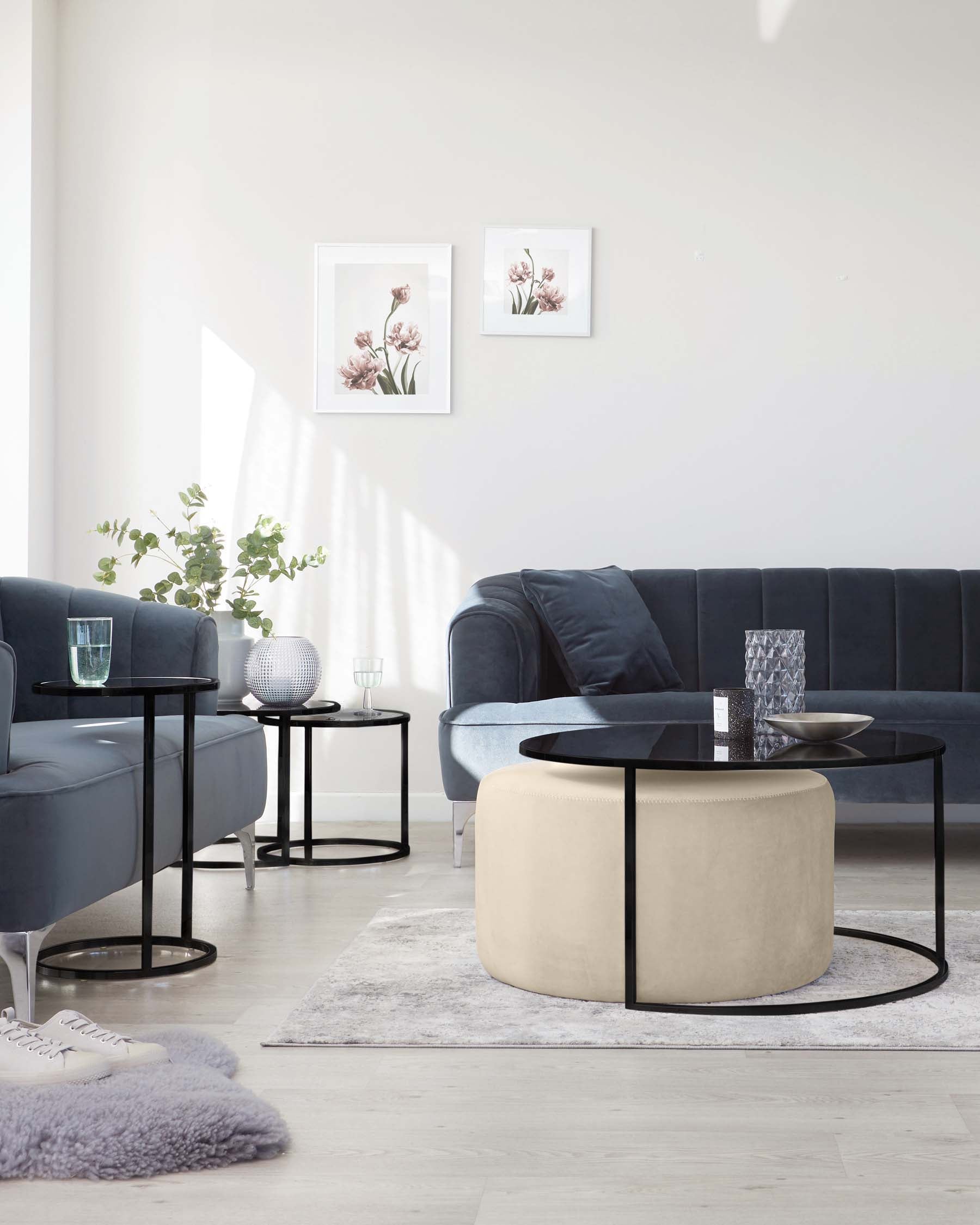 Elegant living room setup featuring a plush navy blue sectional sofa, two round nested side tables with black legs and glass tops, a round coffee table with a black frame and cream-colored upholstered top, and a decorative grey shag rug. The room enjoys soft natural lighting with minimalist wall art and plants enhancing the serene atmosphere.