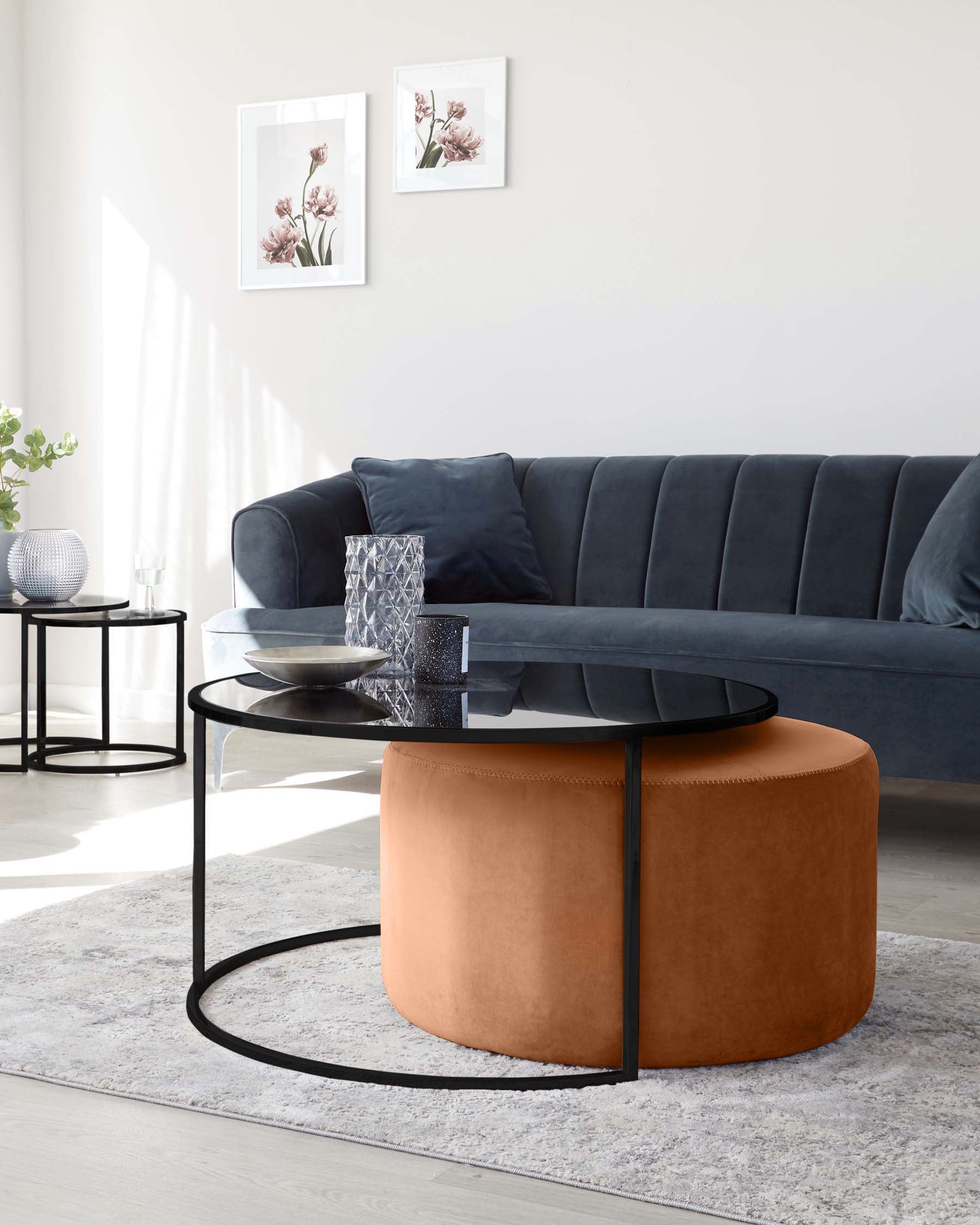 Elegant modern living room furniture including a deep blue velvet sofa with vertical channel tufting and matching pillows, a round black coffee table with a glass top resting on a unique metal base, and a round caramel-coloured leather pouf with visible stitching detail. The setting is complemented with a soft grey textured area rug and minimalist decorative items on the table.