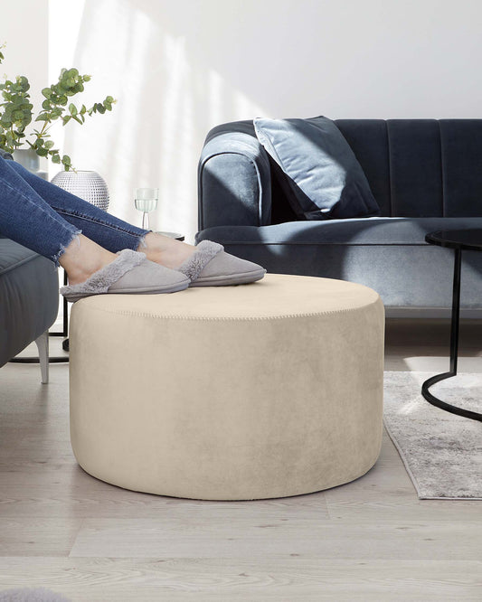 A contemporary living room featuring a luxurious dark blue velvet sofa with plush cushions, complemented by a round, beige upholstered ottoman in the foreground. A sleek black round coffee table is positioned to the side, and the scene is accented by a modern white floor lamp and an indoor potted plant. The space is unified by a soft grey area rug, creating an inviting and stylish atmosphere.