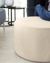 A contemporary round beige ottoman with detailed stitching, positioned in front of a sleek, round black side table with a thin metal frame. A navy blue upholstered chair with a curved silhouette is partially visible in the background. The setting is complemented by a wooden floor.