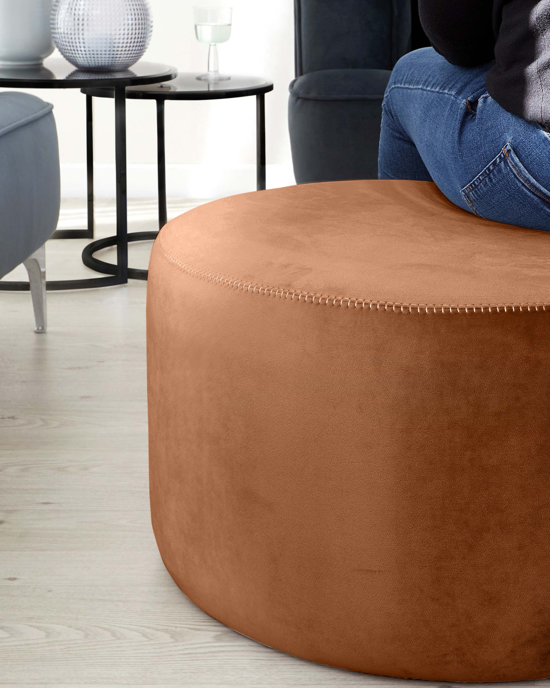 A contemporary living room setting featuring a round caramel-coloured ottoman with detailed stitching on the sides, a sleek black round side table with a textured black and white vase on top, and a portion of a navy upholstered armchair visible on the right.