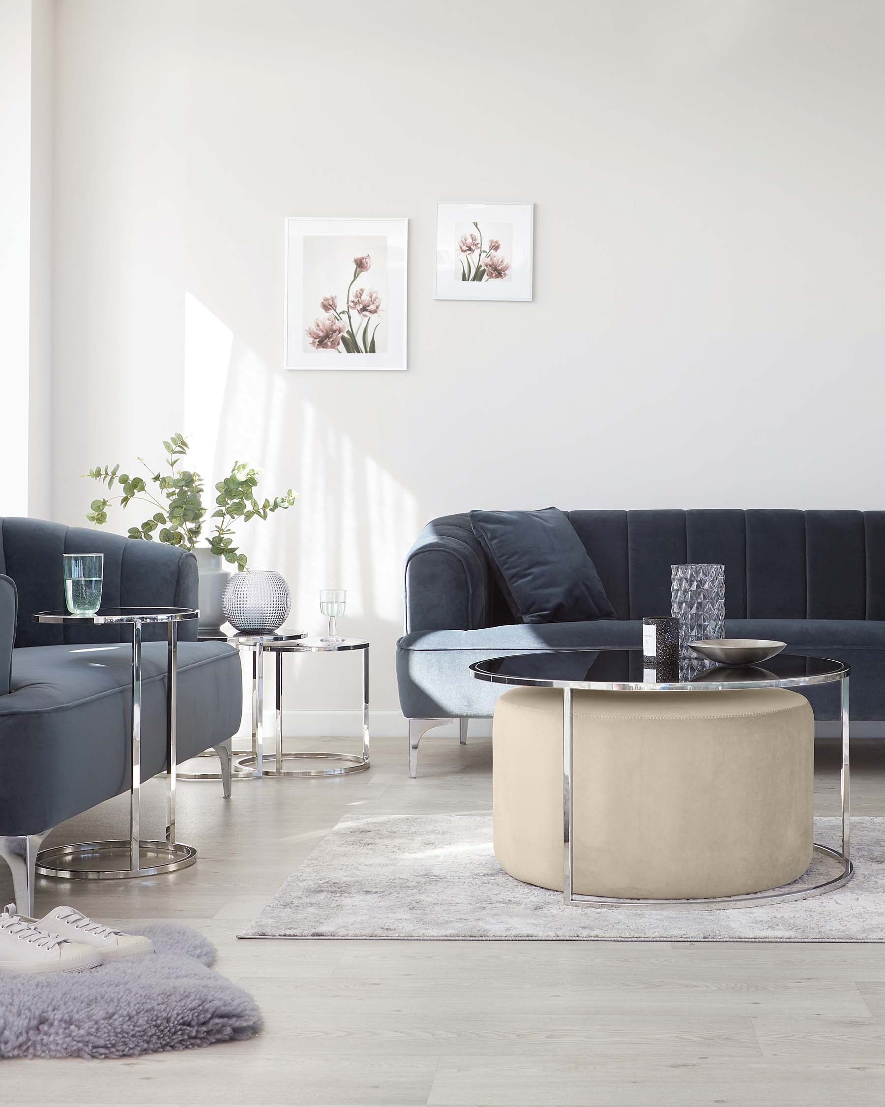 Modern living room furniture arrangement featuring a dark blue velvet upholstered sectional sofa with clean lines and plush cushions, accompanied by two contemporary glass-top end tables with polished chrome frames. A large round coffee table with a reflective glass top is centrally placed, featuring a unique beige ottoman nesting underneath with a soft, textured finish. The ensemble is set against a light hardwood floor and a grey area rug with a soft, plush texture.