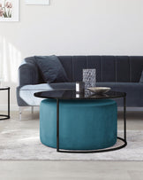 Modern living room furniture arrangement featuring a plush navy blue velvet sofa with back-cushions, accompanied by a round, dark-toned coffee table with a sleek black metal frame and a glistening glass top, which is paired with a teal velvet circular ottoman nestled partially underneath. A smaller black metal side table with a round top is seen to the side. The furniture rests on a textured light grey area rug.