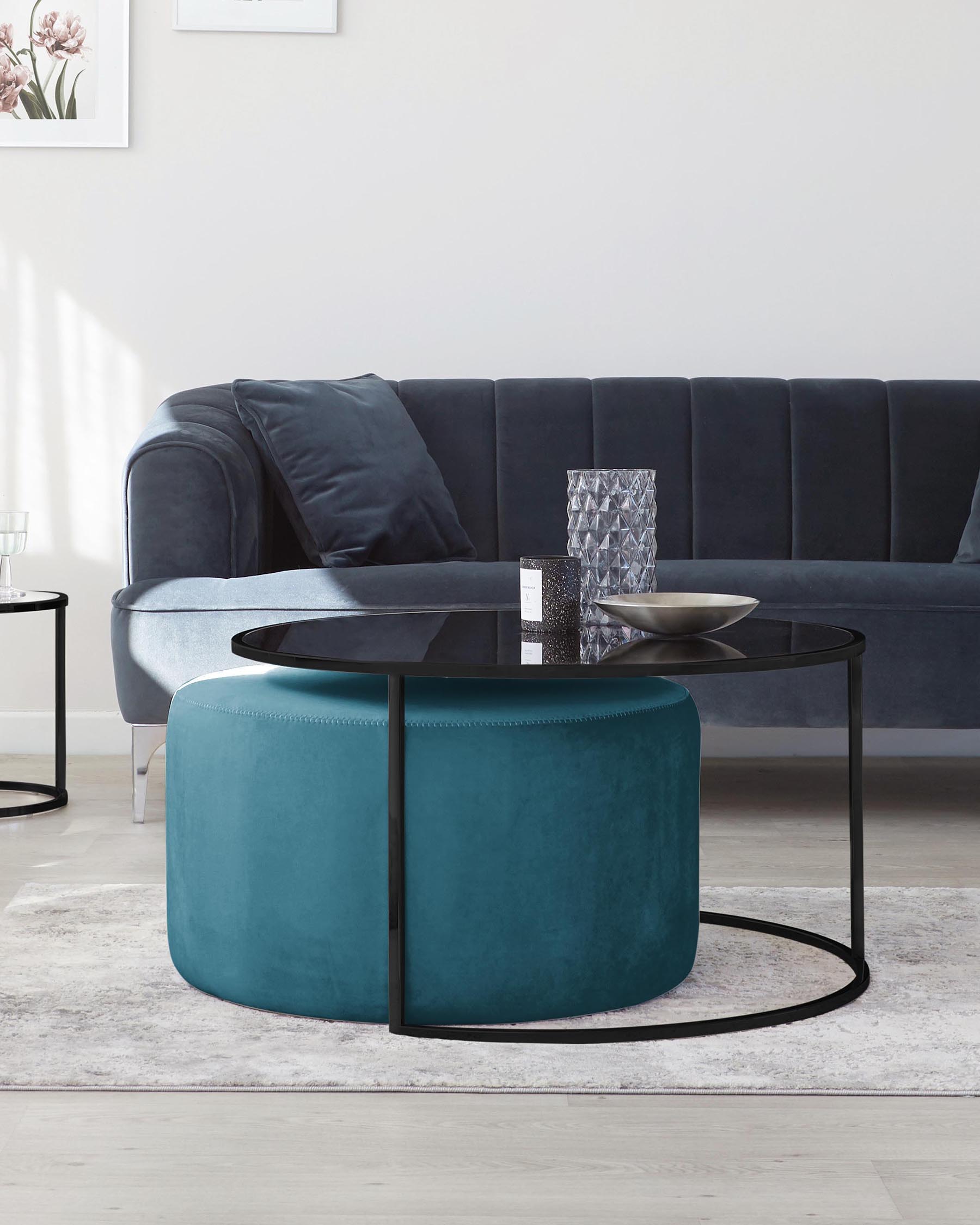 Modern living room featuring a navy blue velvet sofa with plush cushions, complemented by a round teal upholstered ottoman and a circular black metal coffee table with a glass top. The elegant setup is grounded by a soft, light patterned area rug.