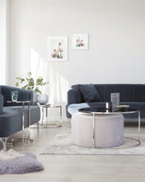 A modern minimalist living room featuring a sleek navy blue velvet sofa with tufted backrest, paired with round glass-top side tables with metallic silver bases. In the centre, a unique circular coffee table with a glass top and a duo of semi-circular white ottomans nested underneath, all atop a soft, light grey area rug.