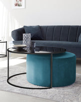 Elegant living room scene featuring a dark grey velvet sofa with vertical tufting and plush cushions. In front, a modern round coffee table with a black metal frame and glass top, accompanied by a blue velvet ottoman with a stitched detailing. The setting is completed by decorative vases on the table, a light grey shaggy area rug beneath, and a simplistic flower painting on the wall.