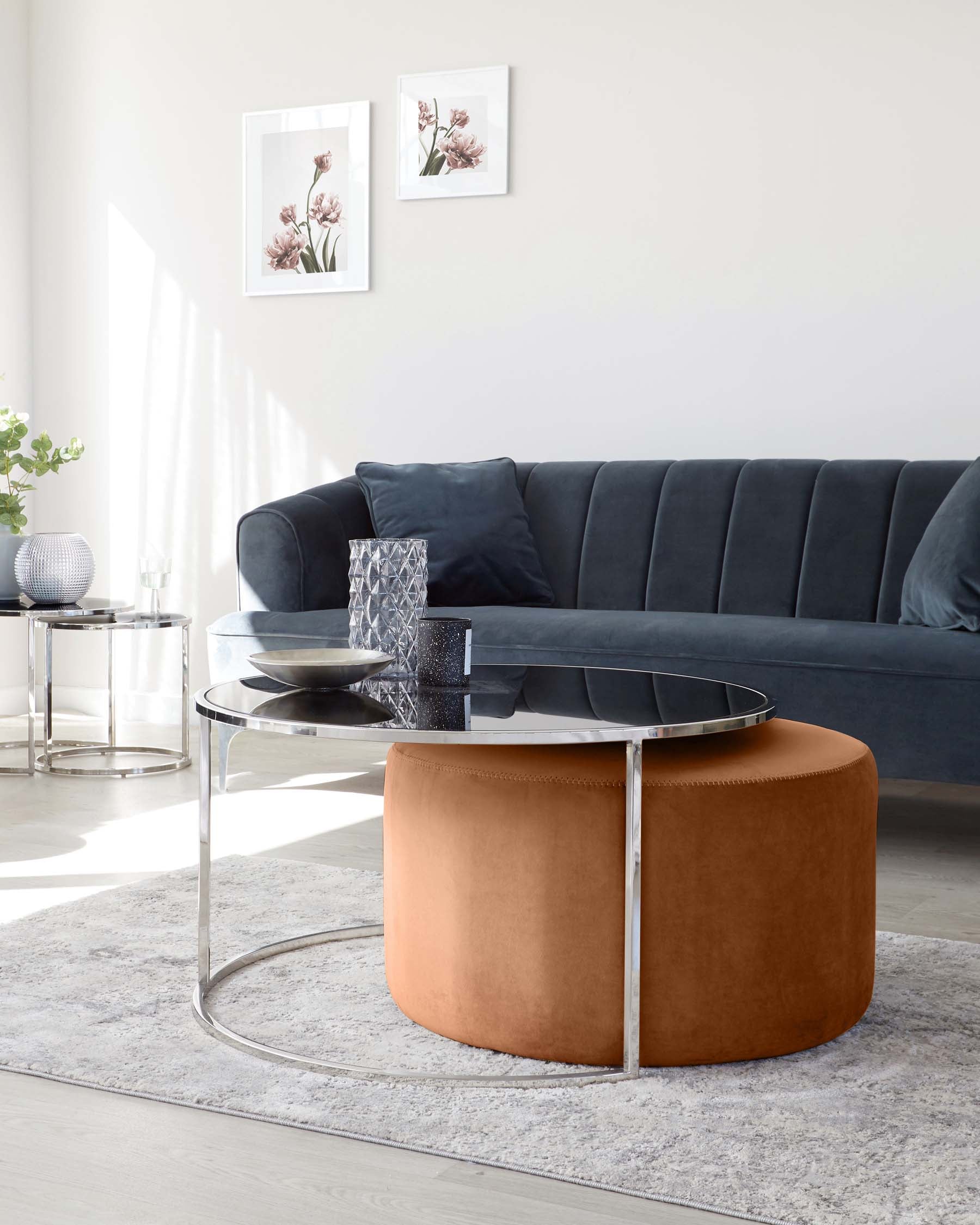 A sleek modern living room featuring a dark grey upholstered sofa with vertical channel tufting, a round glass top coffee table with a unique silver metallic base encircling a cylindrical caramel-coloured ottoman. The setup is complemented by a soft grey area rug.