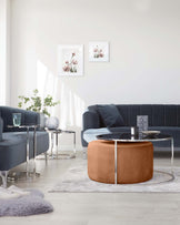 Elegant contemporary living room furniture arrangement featuring a dark blue velvet upholstered sofa, round leather ottoman in caramel tone, a pair of modern glass-top side tables with silver metal frames, and a complementing glass-top coffee table with a unique round design and silver metal base. The room is accented with a plush grey area rug and soft furry throw.