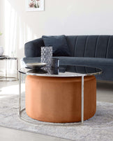 Contemporary living room furniture, featuring a dark grey velvet sofa with plush cushions and a round, tan leather ottoman with metallic silver base. In front of the sofa is a modern round coffee table with a glass top and silver frame.