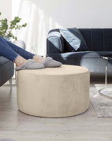 Contemporary beige round ottoman with a velvet-like texture in a modern living room setting, complemented by a plush navy blue sofa with matching velvet cushions, a clear glass side table, and a scatter rug with a muted colour palette on a wooden floor.