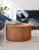 A caramel-coloured round ottoman with a velvety texture is prominently displayed. In the background, there is a dark blue velvet sofa accented by a plush blue pillow. A circular glass side table and a part of a grey area rug are also visible on a light wooden floor.