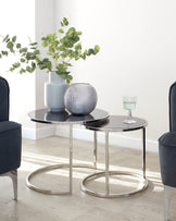 Modern round nesting coffee tables with glossy black tabletops and silver metal frames, alongside two dark upholstered chairs with matching silver legs, set on a light wooden floor.