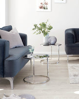 Elegant modern living room furniture featuring a dark blue velvet sofa with metal legs, complemented by a matching armchair. A chic, round, metal-framed glass side table occupies the space between them.