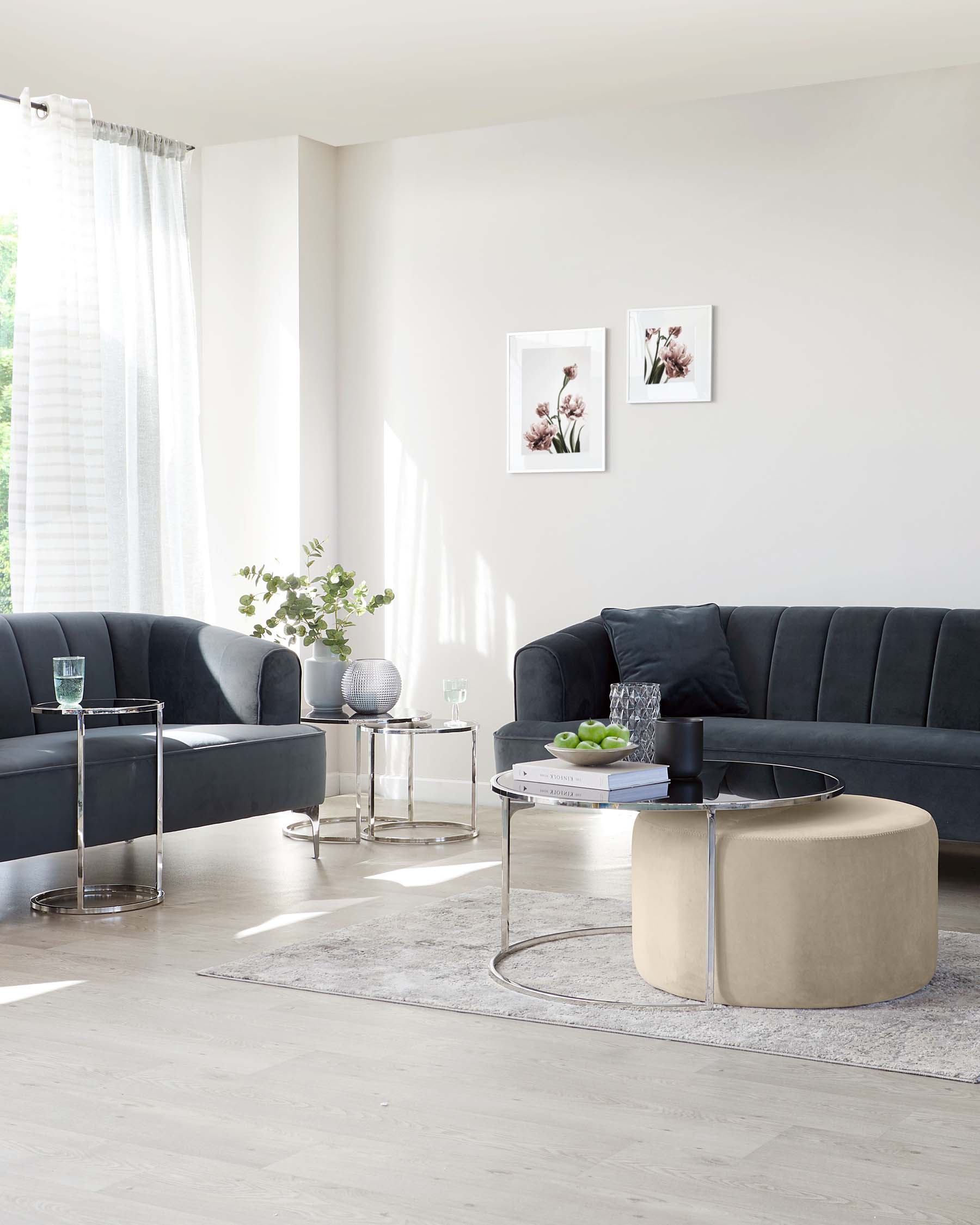 Modern living room setup featuring a dark grey velvet sectional sofa with deep button tufting, accompanied by a circular beige ottoman with a plush finish. A sleek glass-top round coffee table with a metallic base and two matching glass-top side tables provide contemporary accents. The room is completed with a light grey area rug underfoot.