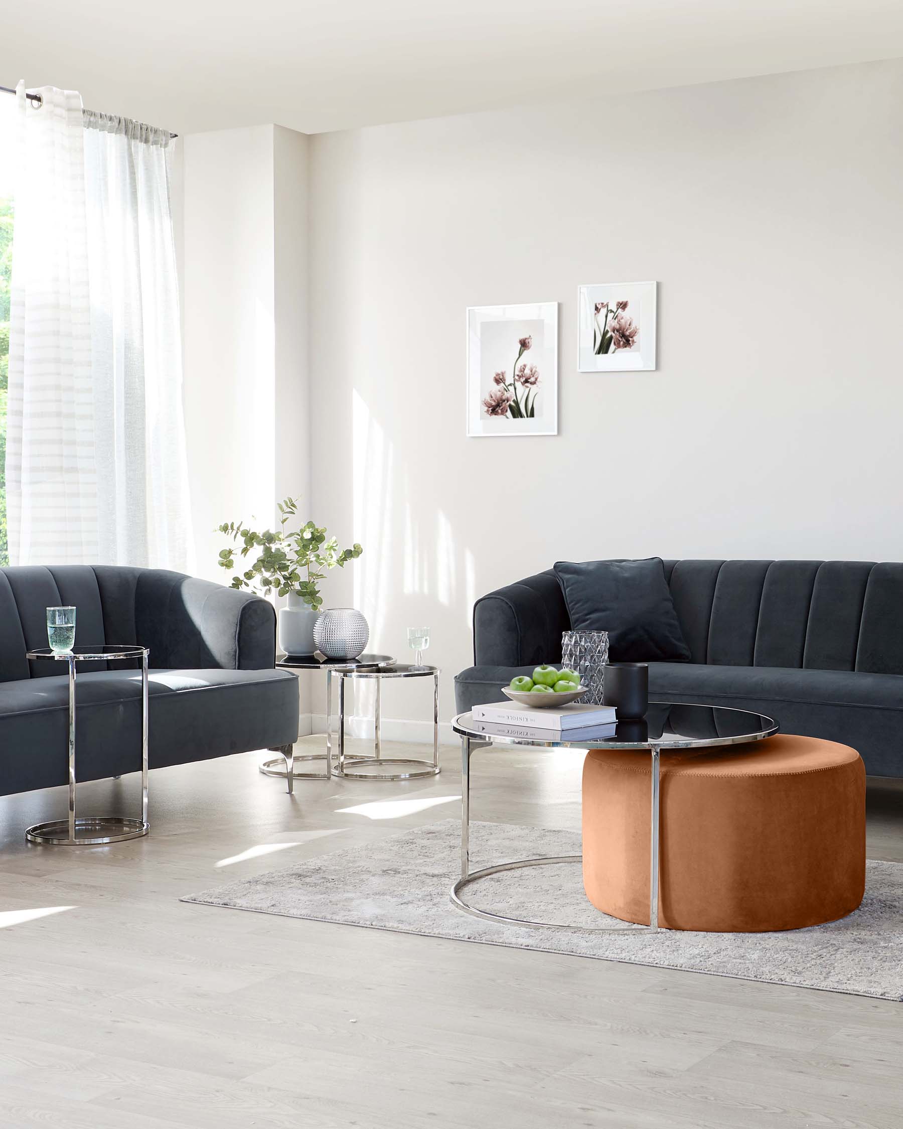 Modern living room featuring a sleek, dark grey sectional sofa with a velvety texture and tufted backrest. In the centre, a unique, two-tiered round coffee table combines a glass top and a caramel-coloured leather bottom tier, supported by a thin, metallic frame. To the side, a matching pair of round, metal-framed side tables with mirrored tops flank the couch, enhancing the room's contemporary elegance. A soft, textured rug anchors the furniture arrangement on a light wooden floor.