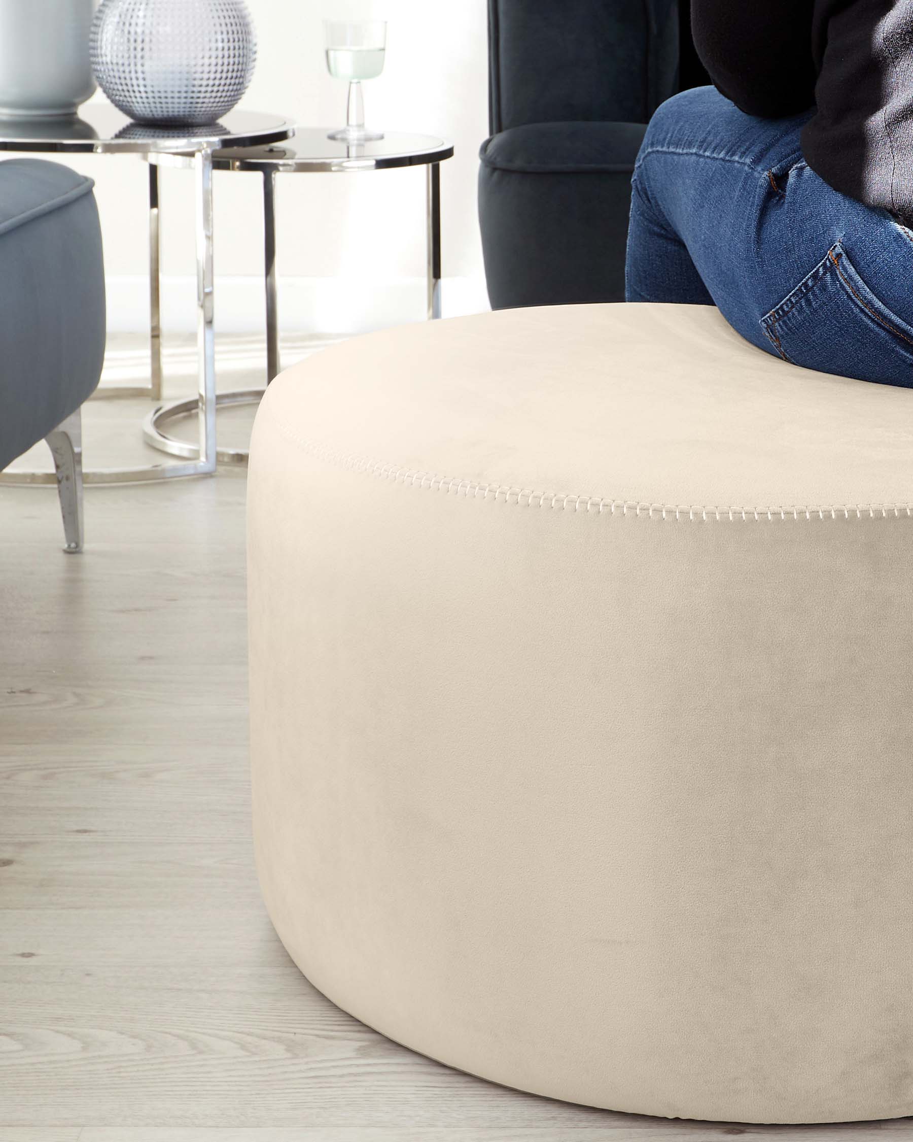 Round beige fabric ottoman with decorative stitching details, near a modern black side table with a glass top, accompanied by a dark blue velvet armchair, all arranged on a light wooden floor with a backdrop of sheer white curtains.