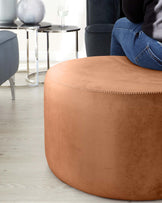 A contemporary round camel-coloured ottoman with decorative stitching is displayed in the foreground, complemented by a sleek black metal and glass side table in the background, which is adjacent to a navy-blue velvet armchair, all staged on a light hardwood floor.