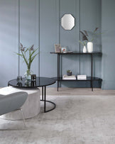 A modern living room featuring a round black coffee table with a sleek metal frame and a smaller accompanying table nested underneath it. Adjacent to the wall is a minimalist black console table with slender legs, hosting an array of decorative items and books. The room is complemented by a soft, grey upholstered ottoman with a metal base, adding a touch of contemporary elegance to the space.