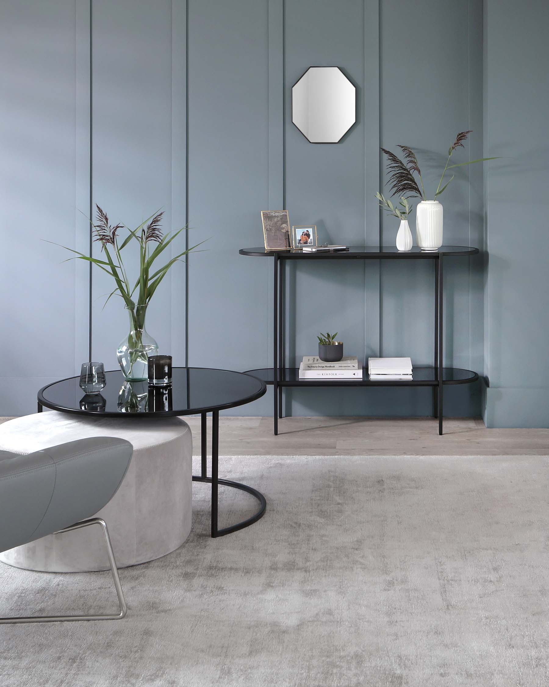 A modern living room featuring a round black coffee table with a sleek metal frame and a smaller accompanying table nested underneath it. Adjacent to the wall is a minimalist black console table with slender legs, hosting an array of decorative items and books. The room is complemented by a soft, grey upholstered ottoman with a metal base, adding a touch of contemporary elegance to the space.