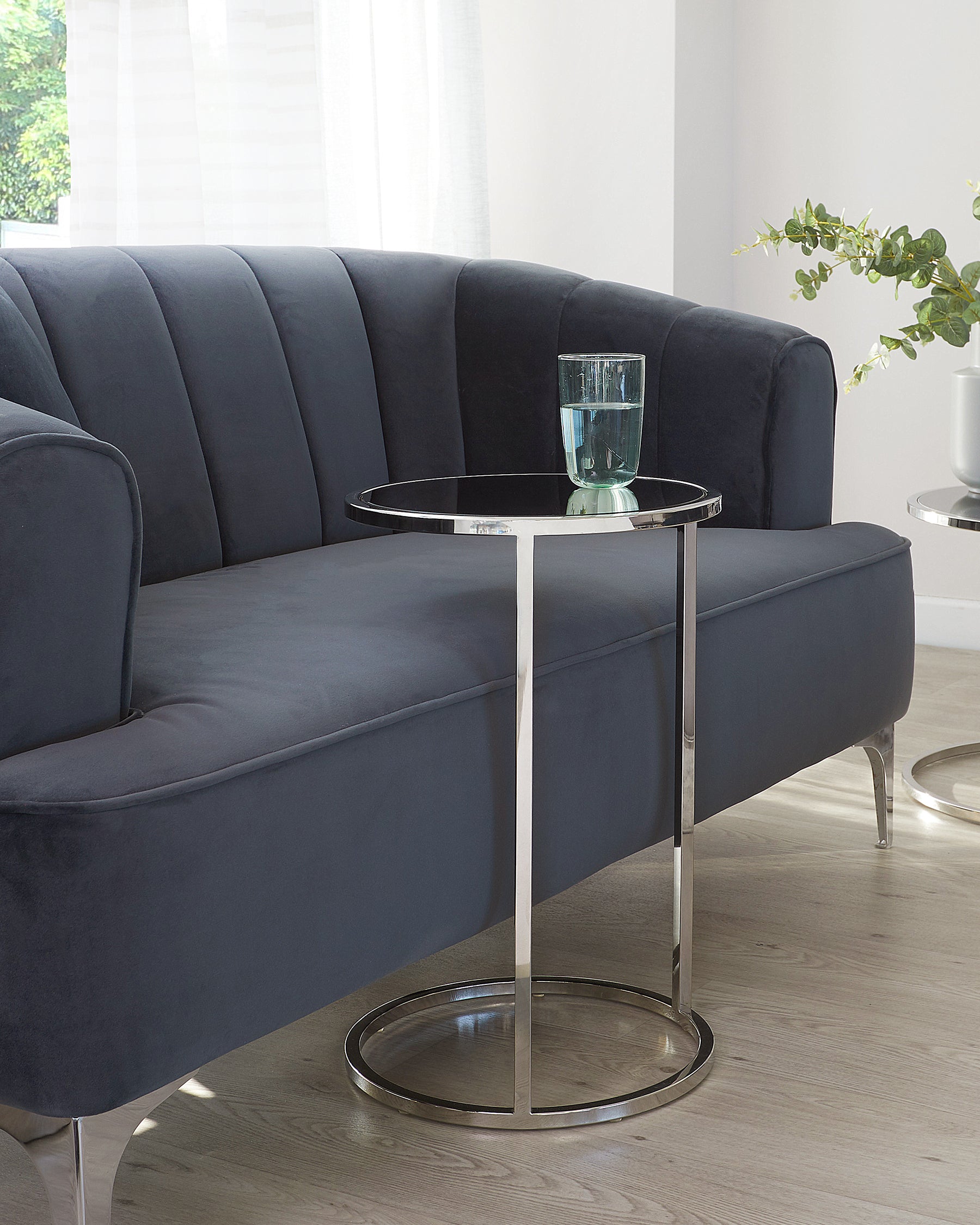 Elegant modern furniture set featuring a curved dark blue velvet sofa with tufted backrest and silver metal legs, accompanied by a minimalist round side table with a silver metal frame and glass top, holding a clear blue glass.