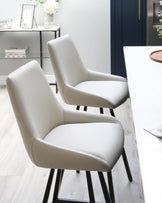 Two modern light grey upholstered dining chairs with a curved backrest and cushioned seat, featuring black metal legs, set against a white minimalist dining table on a light wooden floor, with a shelving unit and decor in the background.