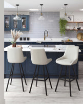 Modern kitchen with two elegant bar stools featuring cream-colored upholstered seats and black tapered legs, positioned at a white countertop island against a backdrop of navy blue cabinetry and subway tile.