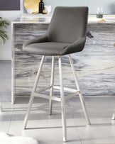 Modern grey upholstered bar stool with a curved backrest and sleek silver metal legs positioned in front of a marble-patterned credenza with decorative accents.