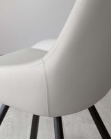 A close-up of a modern chair with a white leather upholstery and detailed stitching along the edges, featuring a curved backrest and seated on slender black metal legs.