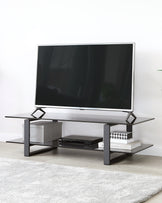 A modern, minimalist TV stand featuring a matte black metal frame with clean lines and two tiers of shelving. The top shelf supports a large flat-screen TV, while the bottom shelf houses electronic equipment and decorative items. The design includes geometric side accents that contribute to its contemporary aesthetic.