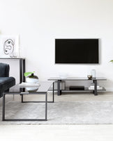 Modern minimalist living room furniture set including a matte black low-profile TV stand with a sleek finish, an adjoining rectangular coffee table with a glass top and metal frame, and a small side table with a matching glass top next to a navy blue upholstered chair. A light grey shaggy area rug anchors the ensemble on a light wooden floor.