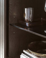 A contemporary dark wood display cabinet with reflective gloss finish, featuring glass shelves and clean lines, partially filled with decorative items and books.