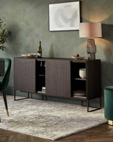 Contemporary sideboard in a dark finish with textured front panels and slim metallic legs, accompanied by an abstract patterned area rug in muted tones. The sideboard features open shelving and is accessorized with decorative items including a table lamp, plates, and a bottle of wine.