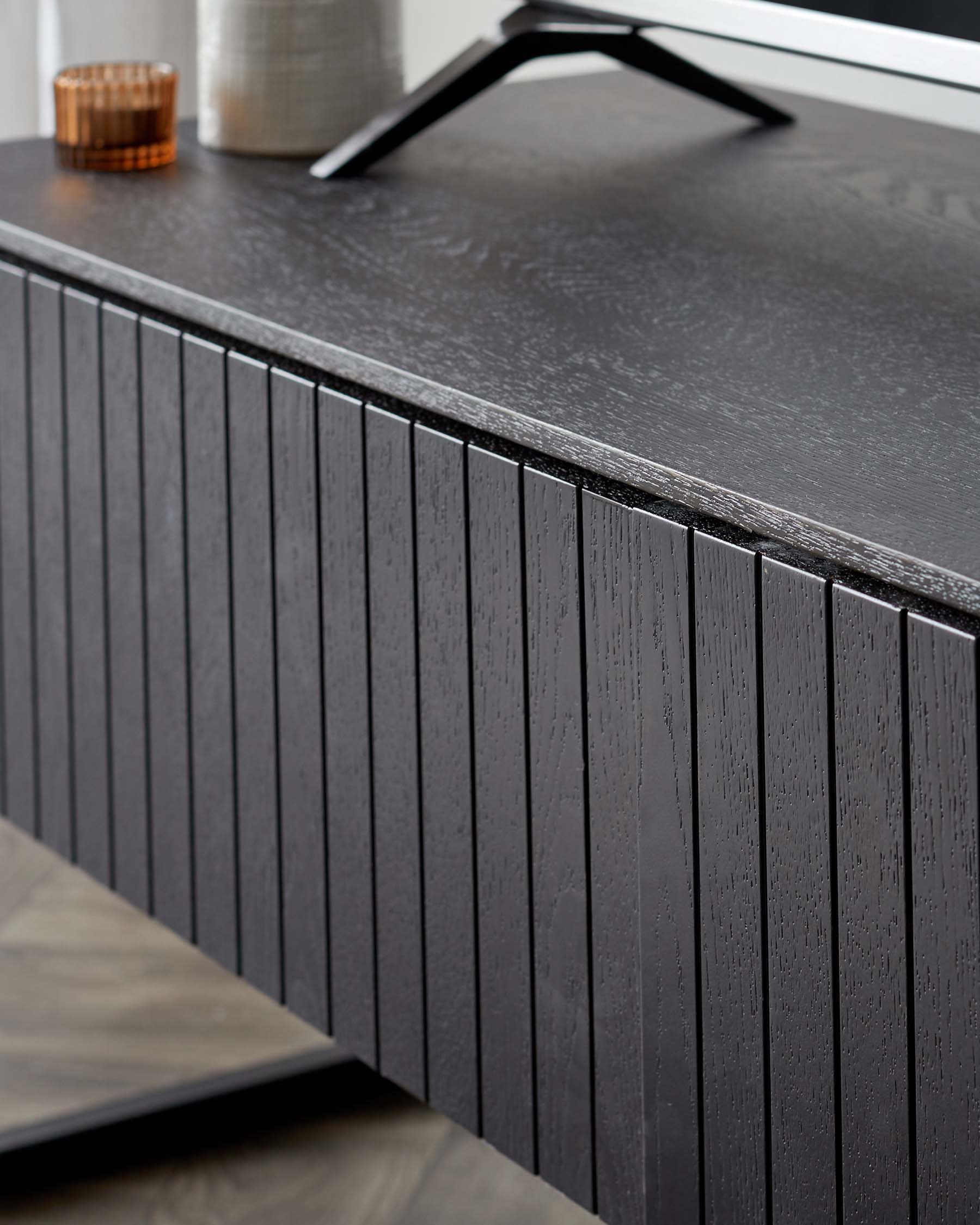 Modern dark wooden sideboard with a textured finish and vertical grooves, featuring a sleek black countertop, paired with a unique matte black metal handle. Decorative items include a small round candle holder and a textured grey vase on top.