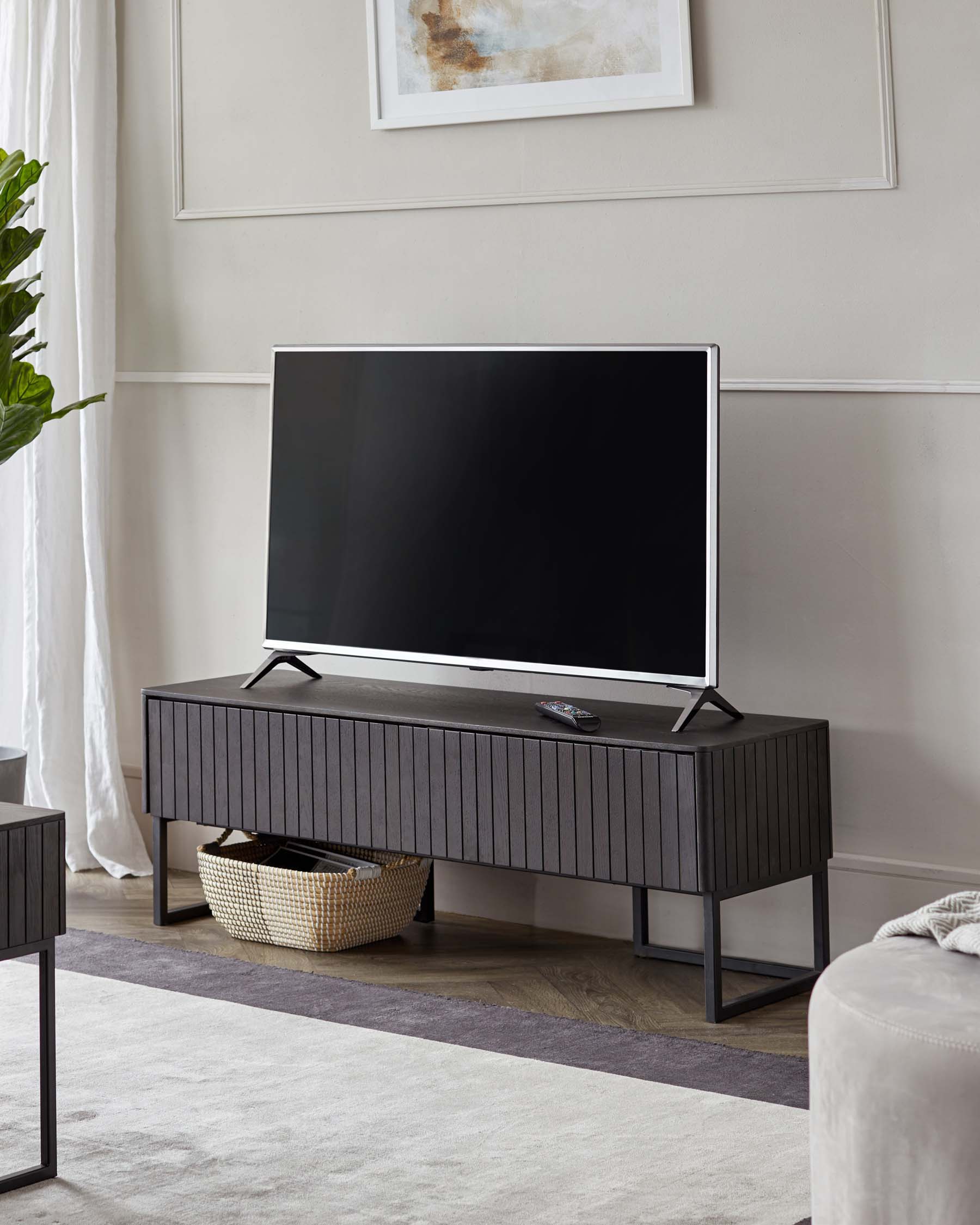 Modern charcoal television stand with a slim rectangular silhouette and textured front, elevated on a minimalist black metal frame. A decorative basket sits beneath the stand on the right.