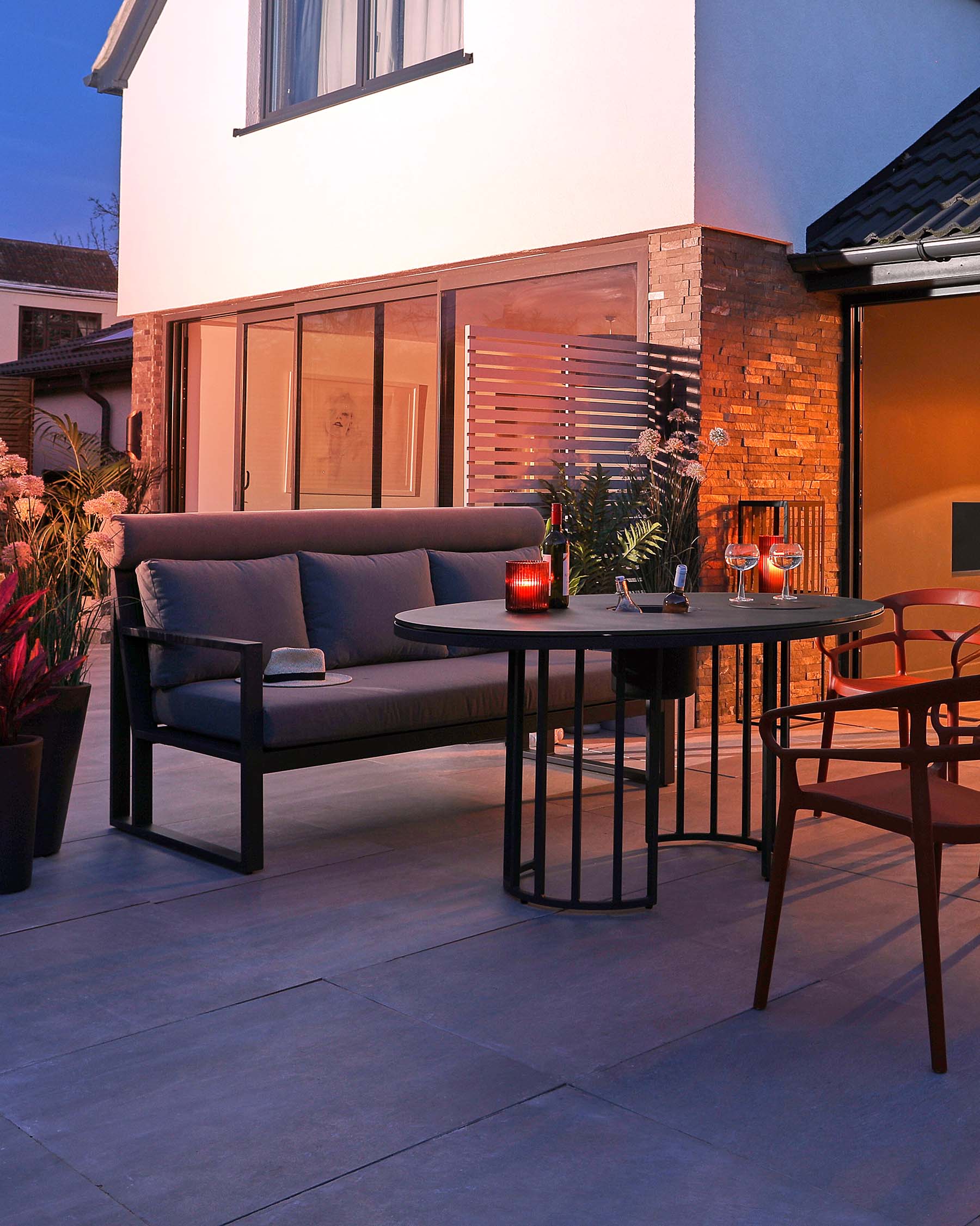 Modern outdoor furniture set on a patio at dusk, including a sleek dark grey two-seater sofa with black metal frame, a round black metal side table with a glass top, and a stylish brown dining chair with a curved back and slender legs.