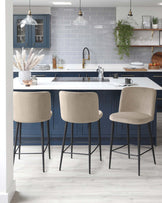 Three modern bar stools with beige upholstery and black metal legs, positioned at a kitchen island with a white countertop and navy blue cabinets.