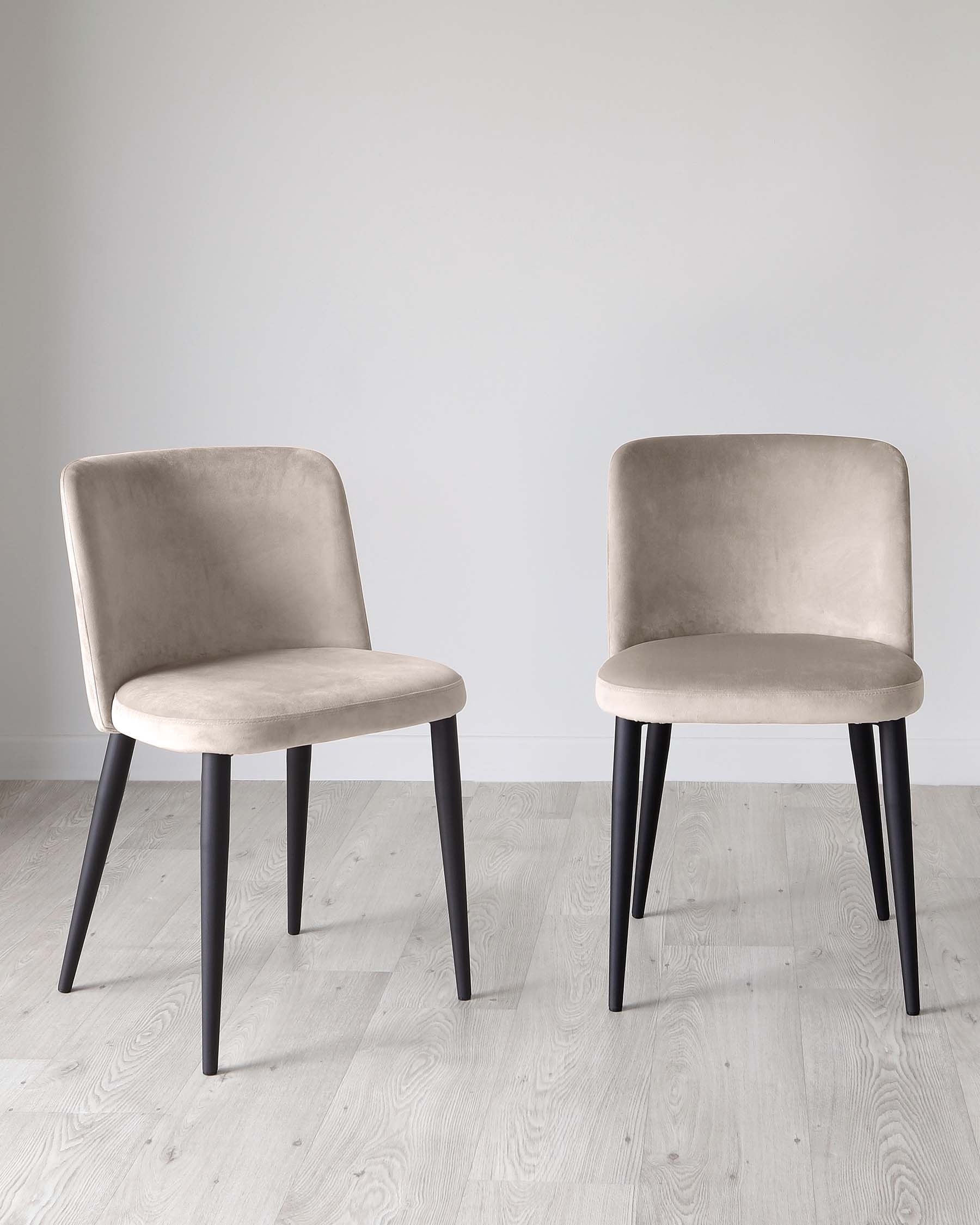 Two contemporary-style dining chairs with velvet taupe upholstery and sleek black legs on a light wood floor against a grey wall.