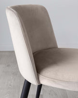 A contemporary dining chair featuring a curved backrest and cushioned seat with light beige upholstery. The chair has visible stitching details along the edges and is supported by tapered black metal legs. The design offers a blend of modern minimalism and comfort.