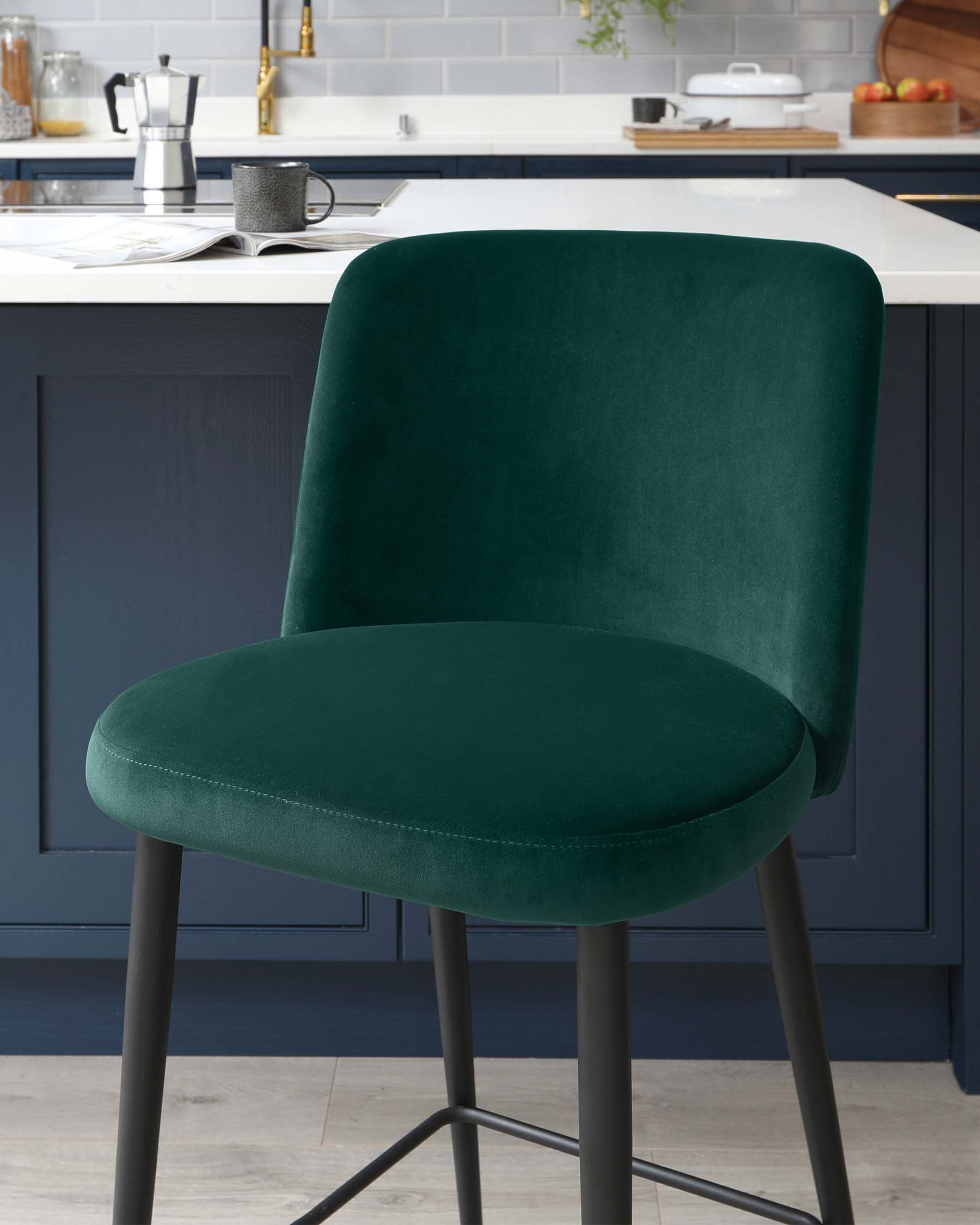 Modern emerald green velvet upholstered bar stool with a curved backrest and black metal legs set against a kitchen island background.